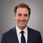 A man with brown hair and wearing a dark blue suit with a white undershirt and dark blue tie smiles against a gray backdrop.