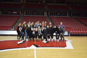 A group of students stands on the center of the basketball court at the Kohl Center on a large motion W logo.