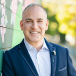 David Grossman, a man with gray hair wearing a navy suit jackey with light blue button up shirt underneath, smiling and leaning against a green wall outside.