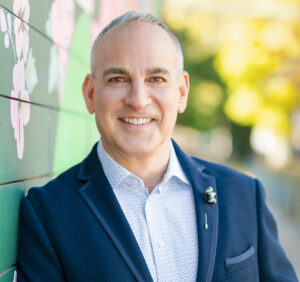 David Grossman, a man with gray hair wearing a navy suit jackey with light blue button up shirt underneath, smiling and leaning against a green wall outside.