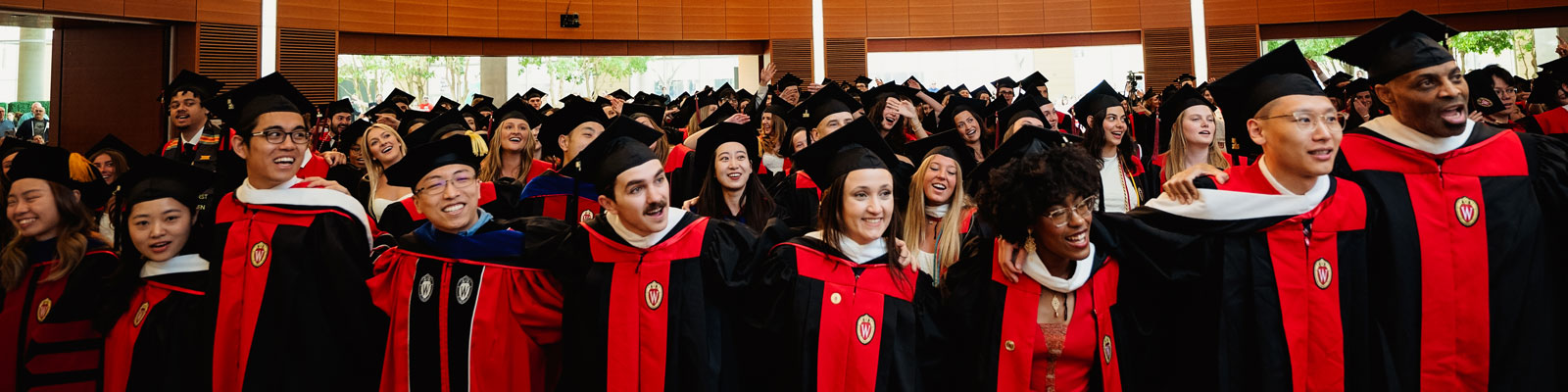 Graduation ceremony with students in black caps and gowns, red and white stoles, indoors with natural light.