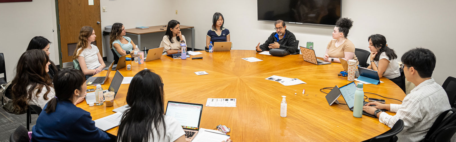 Graduate students sit around a large round wooden table during a research seminar with professor Dhavan Shah.