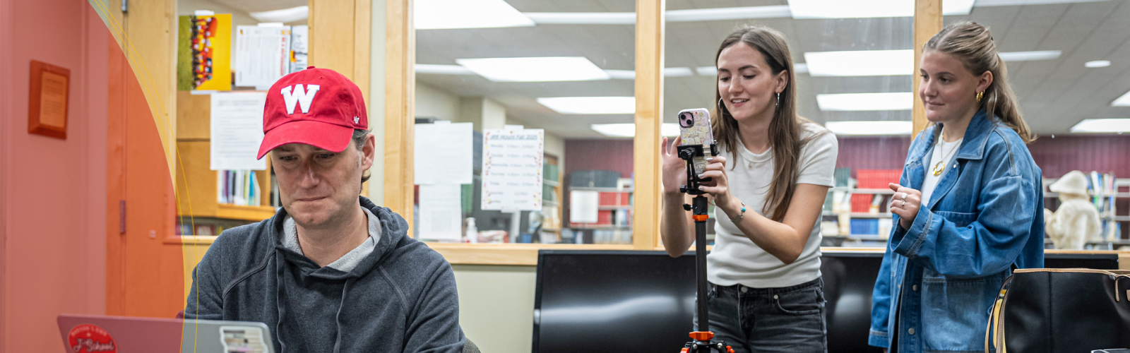 A man wearing a red “W” baseball cap works on a laptop at a table while two young women behind him set up a smartphone on a tripod, smiling as they prepare to record in a classroom or office setting.