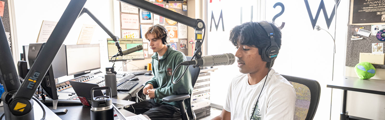 Two students wearing headphones sit in a radio studio, speaking into microphones while operating audio equipment and a computer, with sunlight coming through a window behind them.