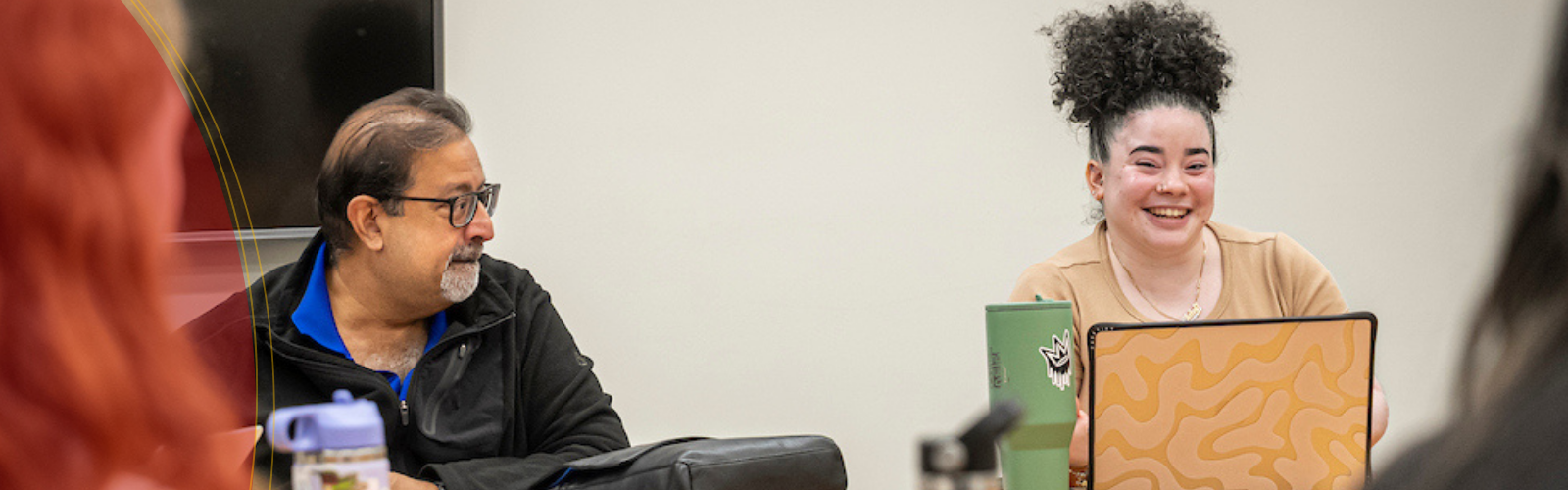 A group sits around a table in a meeting room; a man with glasses looks toward a smiling woman with curly hair as she speaks, with laptops, water bottles, and a large tumbler visible on the table.