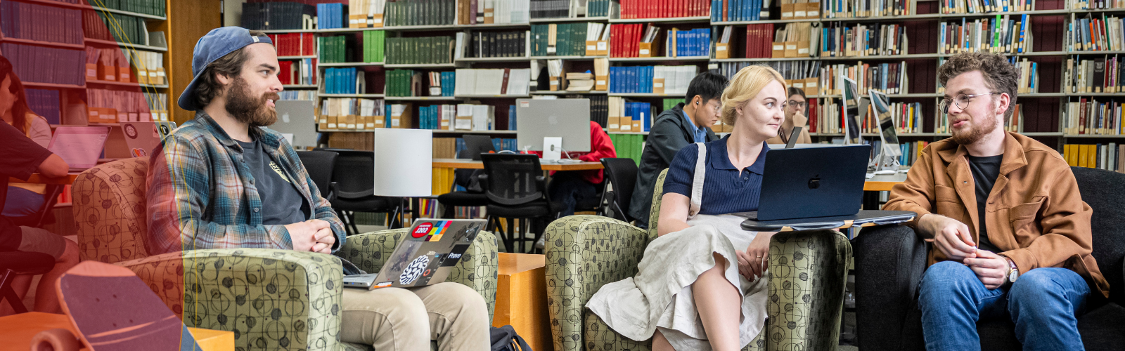 Three students chat in a library; a male student on the right talks while a male and a female student listen.