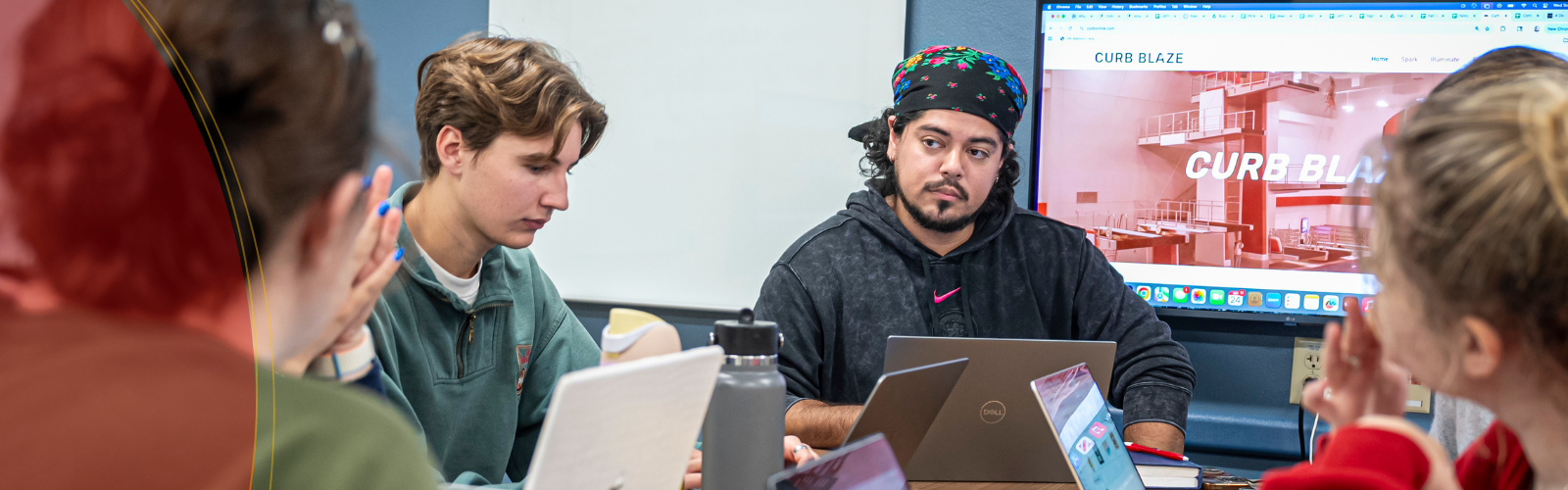 Students collaborate around a table with laptops in a classroom; one student gestures while another looks down at notes, and a screen in the background displays a website labeled “CURB BLAZE.”