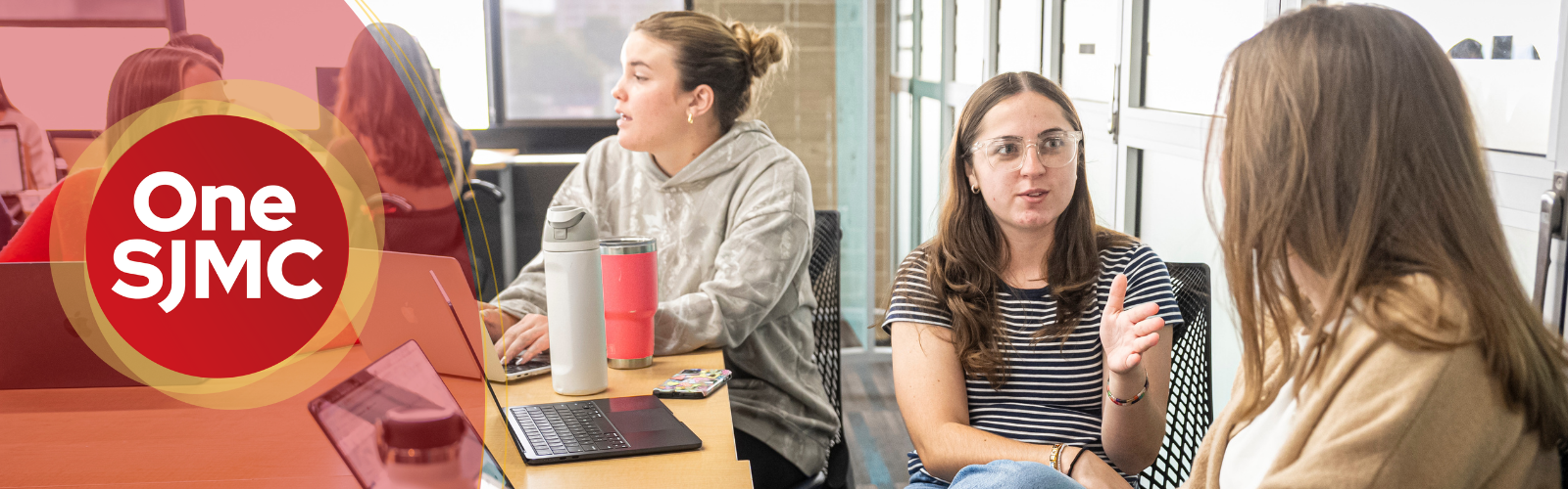 Three college-aged students sit around a table in a bright classroom, engaged in discussion while using laptops; one student gestures as she speaks, while another types and a third listens. A red graphic overlay on the left reads “One SJMC.”