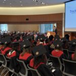 An auditorium filled with graduates watching the keynote speaker at their graduation celebration.