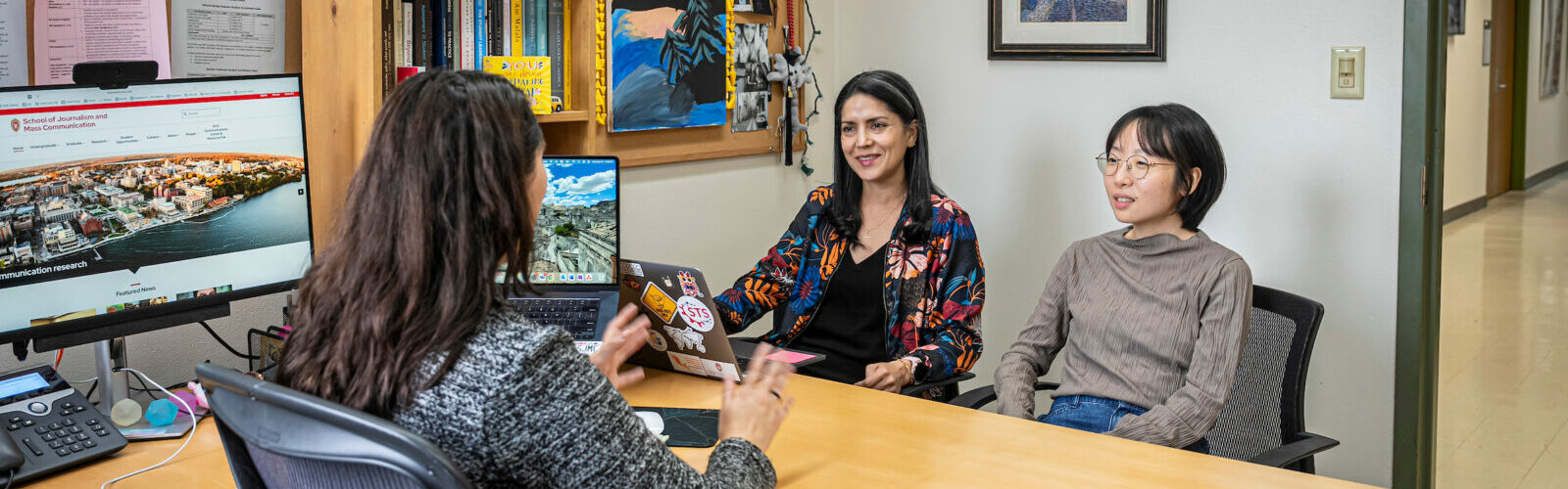An advisor with medium length brown hair sitting at her desk gives advice to two female students sitting across from her.