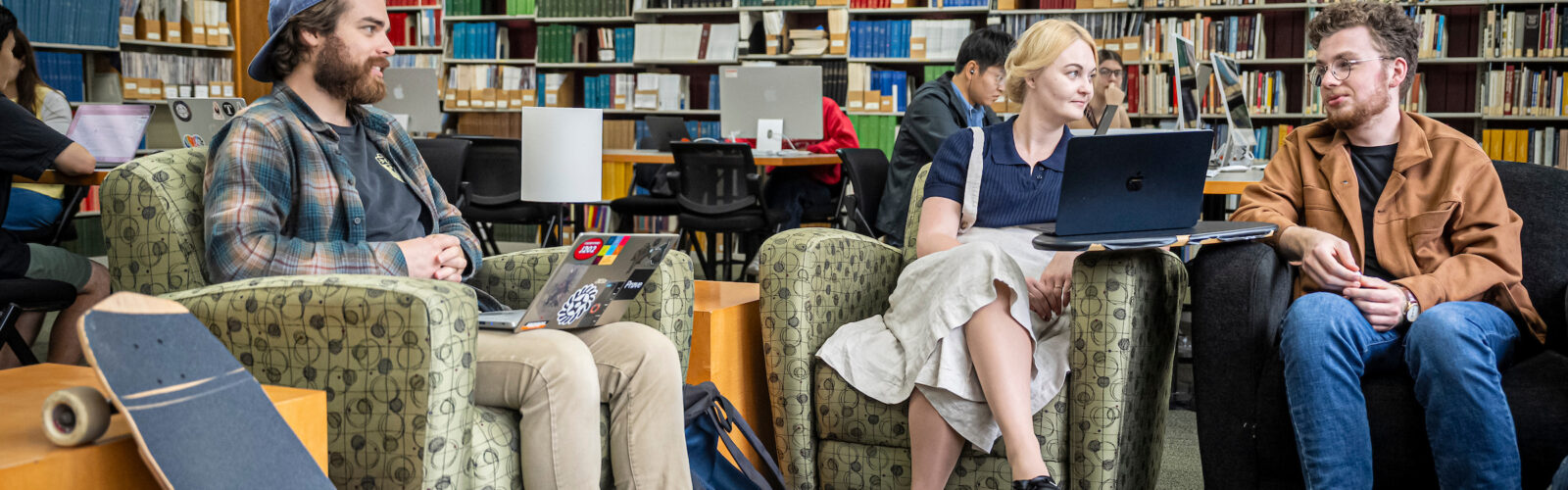 Three students chat in a library; one male student on the far right talks while another male and female student listen.