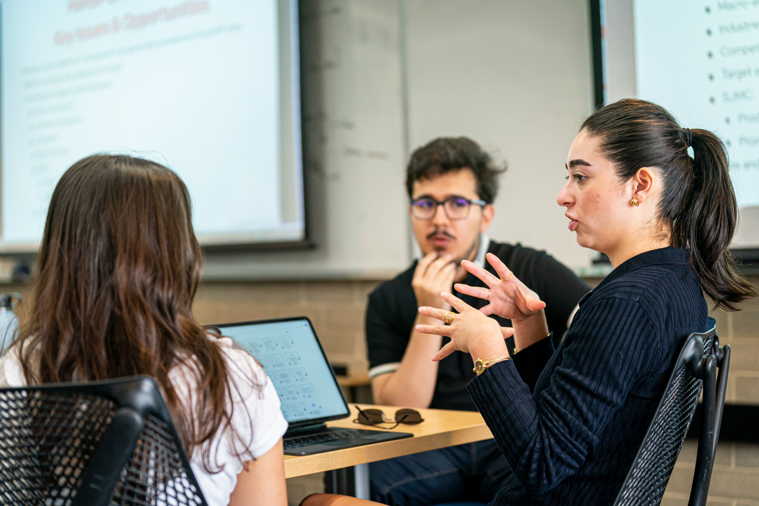 A female student with dark hair and a black sweater leads a discussion with her two classmates.