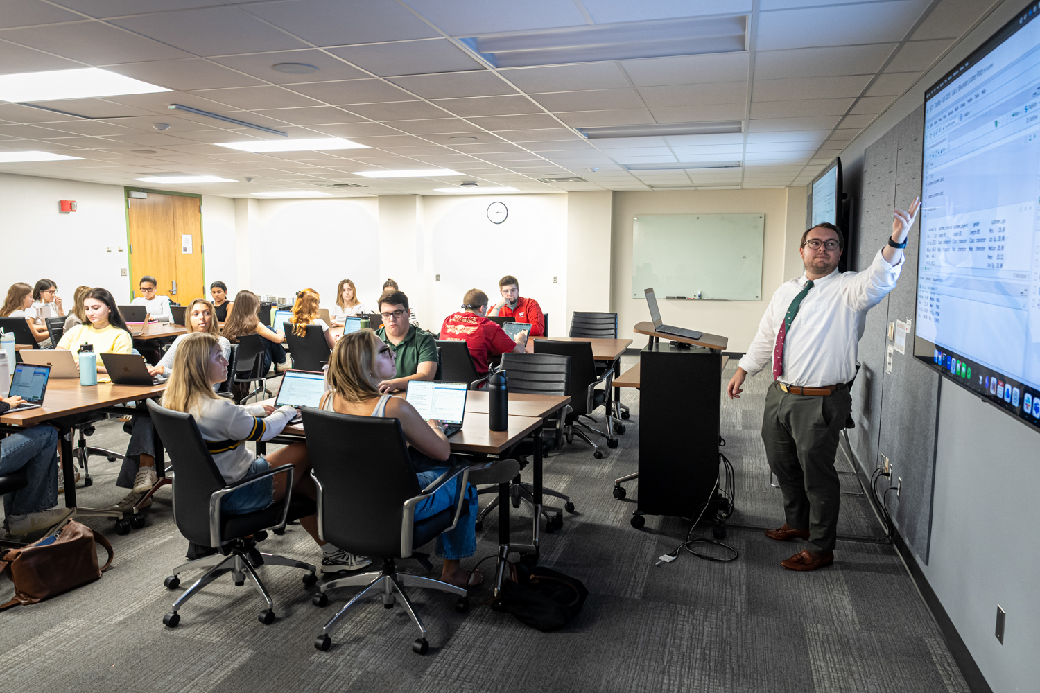 A male teacher in a white shirt and tie leads a course and points to a screen in front of a classroom of students.