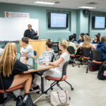 A man with dark hair wearing a black sweatshirt leads a classroom of students