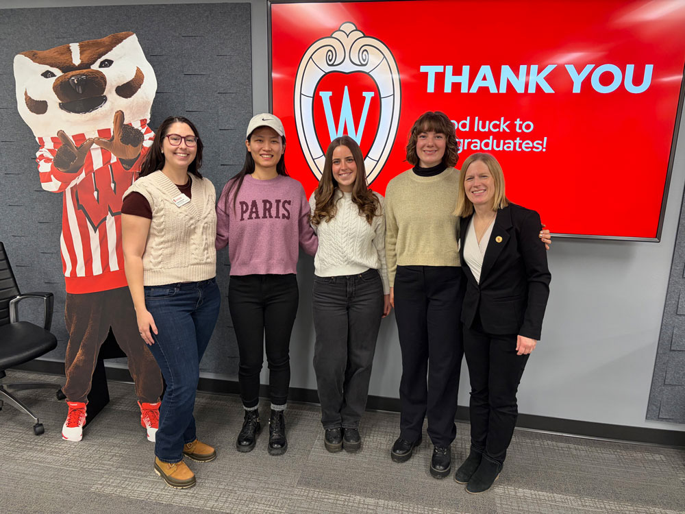 Five women pose together in front of a cardboard cutout of Bucky Badger and red screen that reads "Thank you"