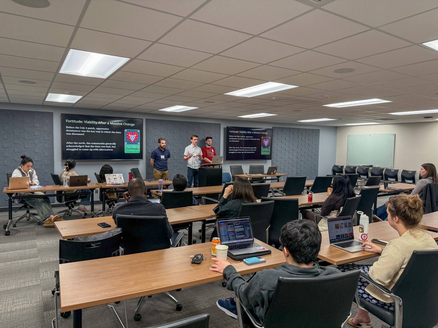 Three male students present their research in front of a classroom of other graduate students.