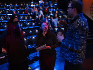 A woman in a black shirt with a headset talks to two other people on a TV set