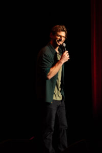 A man wearing glasses and a dark gray shirt with a microphone on stage.