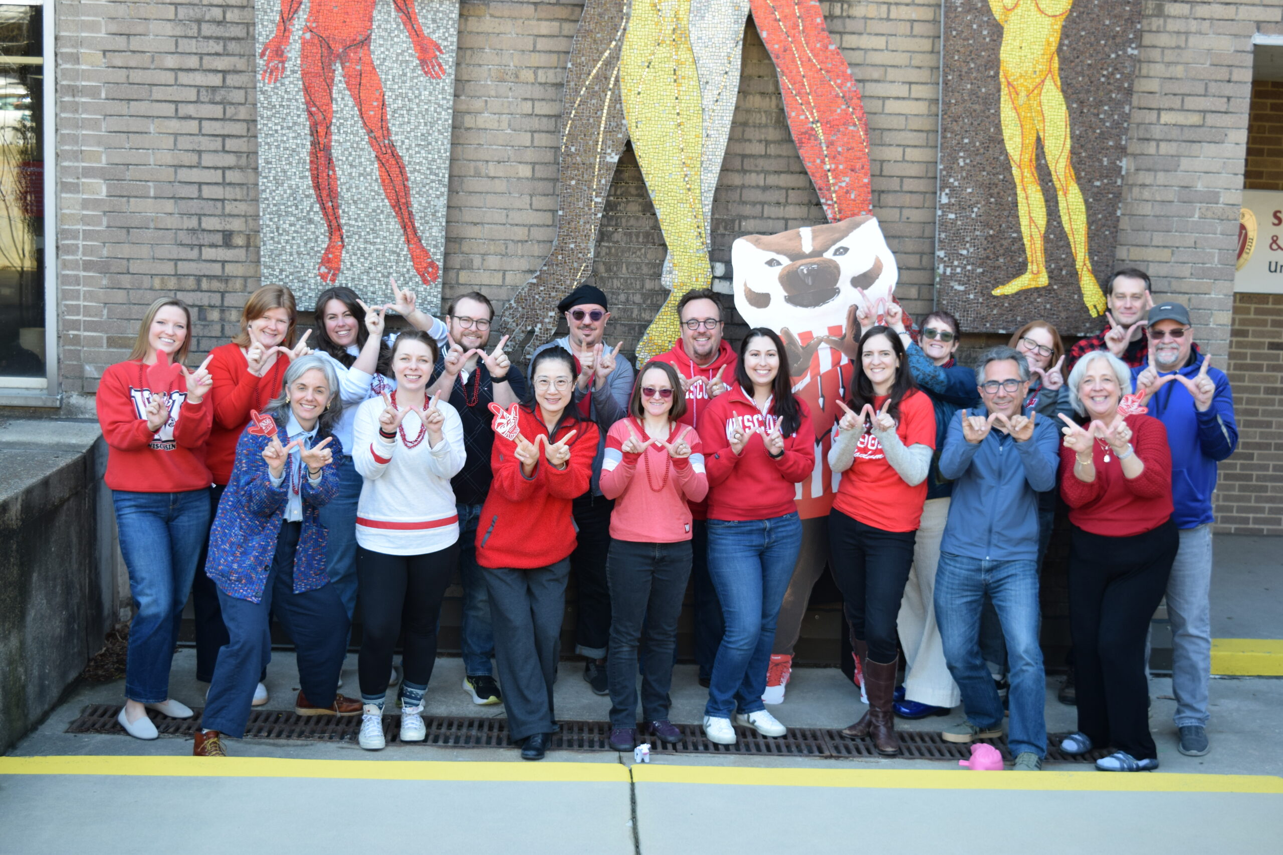 A group of men and women pose outside with a cardboard cutout of Bucky Badger and holding up their hands in a "W" shape