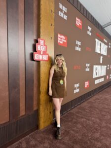 A woman with blond hair wearing a brown dress poses on a brown carpet with a walk behind her that reads "The Height of Kevin Hart"