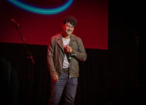 A man wearing a brown jacket and gray jeans holding a microphone on a stage