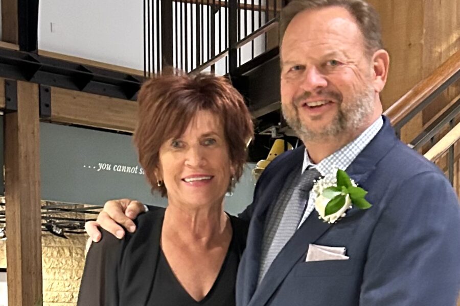 A woman with short brown hair in a black shirt smiles with a man in a dark gray suit.