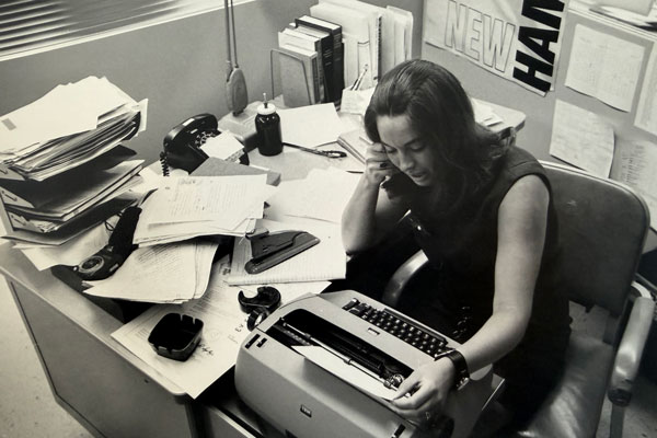 A black and white photo of a woman sitting at a desk covered in papers while on the phone and working on a typewriter.