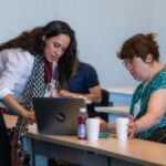 A woman with dark hair and a white shirt assists another woman on her laptop