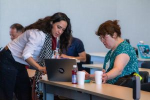A woman with dark hair and a white shirt assists another woman on her laptop