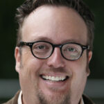 A man with brown hair and glasses smiling.