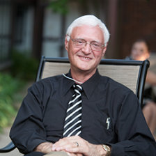A man with white hair and glasses sitting in a chair outside and wearing a black collared shirt and striped tie.