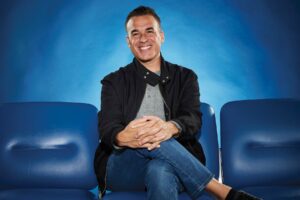 A man with dark hair, wearing a gray shirt with a black jacket and jeans, sits smiling with his legs crossed between blue auditorium seats.