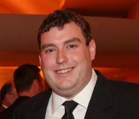 A man with brown hair, smiling and wearing a black suit with a white shirt and black tie.