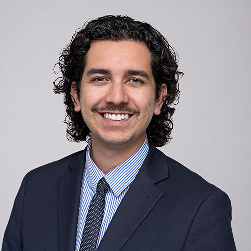 Professional headshot of a smiling man with curly dark hair and a mustache, wearing a navy suit, blue-and-white striped shirt, and patterned tie against a light gray background.
