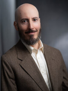 Portrait of a bald, bearded man wearing a brown blazer and white shirt, facing the camera against a dark gray background.