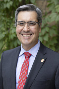A man with dark hair and glasses smiling outside in a black suit jacket with a blue shirt and red pattered tie