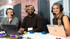 Three radio hosts wearing headsets and microphones smile at a table with laptops in an ESPN Radio studio decorated with a brick wall, black shutters and purple-and-gold Mardi Gras beads.